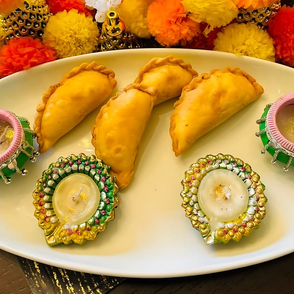 Four golden-brown Indian pastries on a white plate, surrounded by decorative candles and colorful flowers.