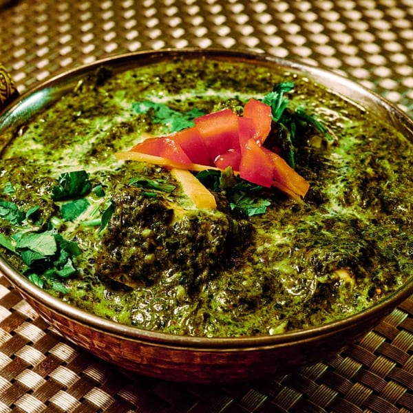 A copper bowl of vibrant green Indian palak paneer, garnished with red tomatoes and fresh cilantro.