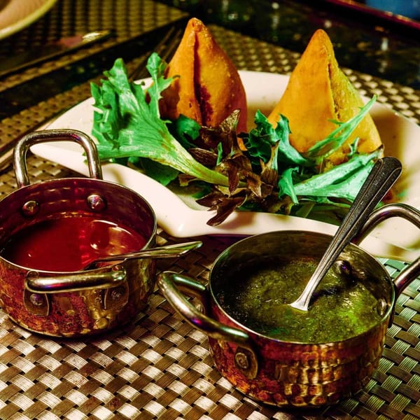 Two golden samosas with fresh greens, served with red and green chutneys in small copper pots.